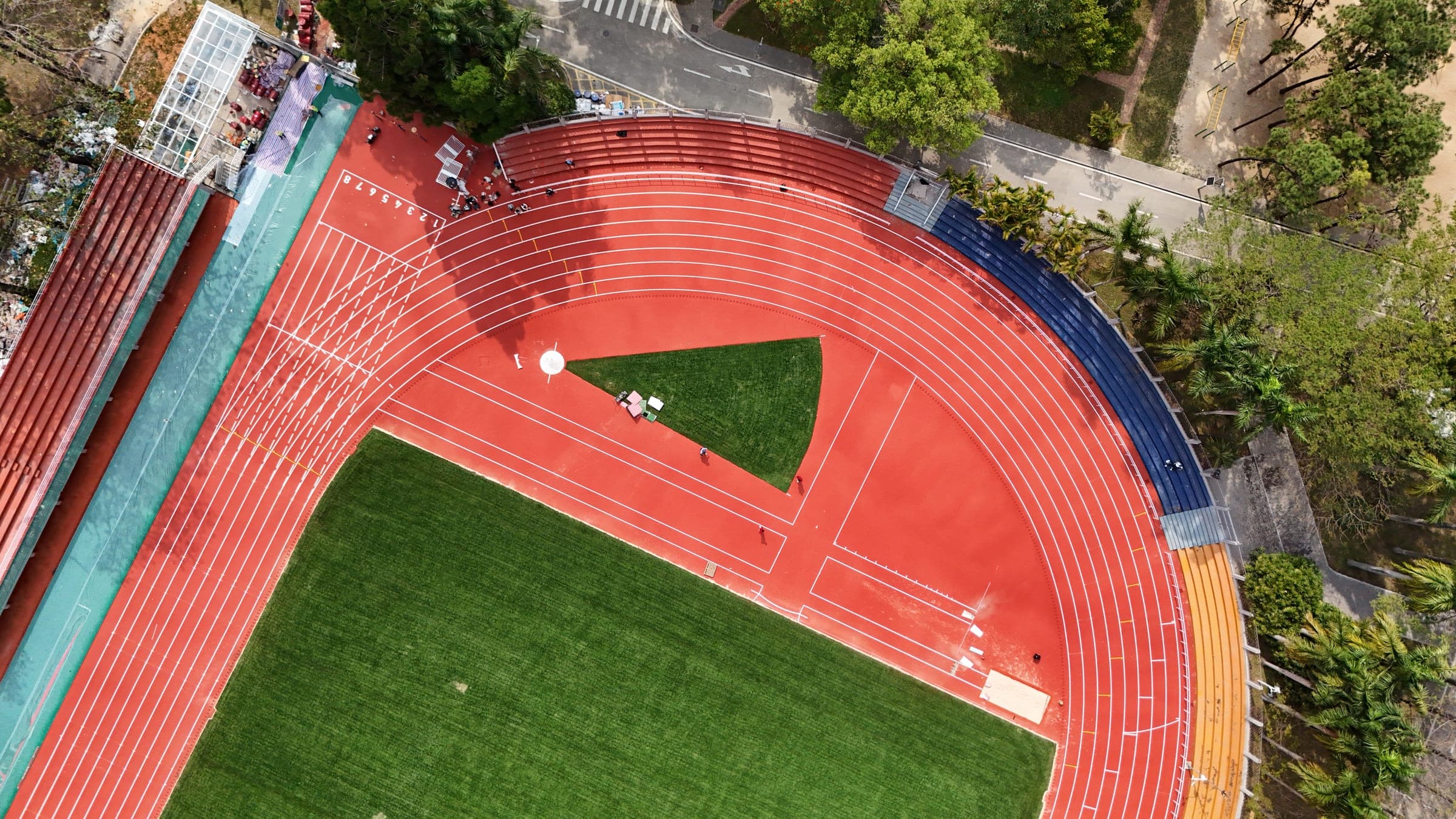 The Guangzhou training stadium from directly overhead, the red track curving around a green infield with blue and orange seating banks along the outside and dense trees beyond.